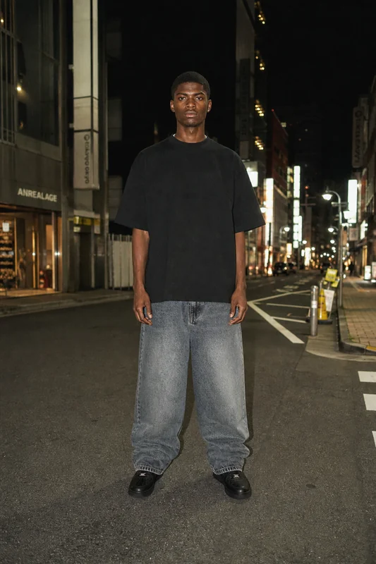 Tall man stands in the middle of a city street at night, wearing a black oversized T-shirt, baggy gray jeans, and black shoes.