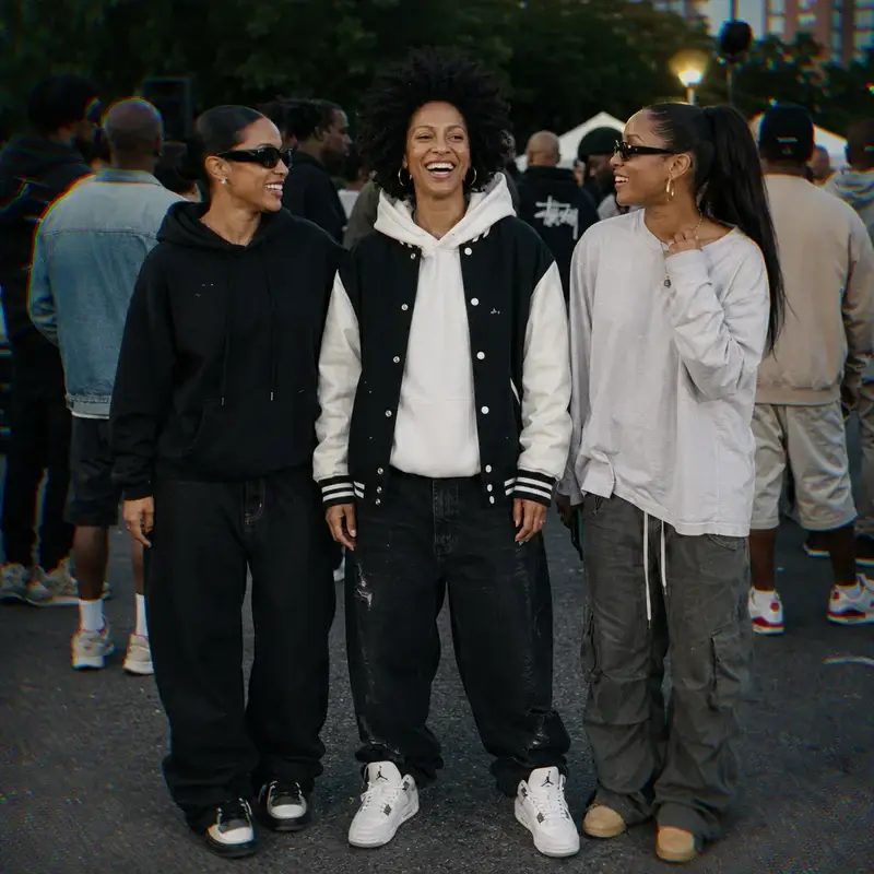 Three friends laughing together at an outdoor street event, with the center person wearing an afro and a varsity jacket over a white hoodie.