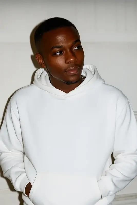 Young man in a white hoodie looking to his left against a light background, hands in pockets.
